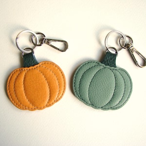 May include: Two pumpkin-shaped keyrings. One is orange, the other is green. Both have a dark green leather loop and silver-coloured metal hardware. The keyrings are made of stitched leather and are designed to resemble pumpkins.