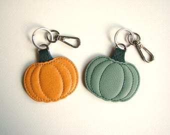 May include: Two pumpkin-shaped keychains. One is orange, the other is green. Both have a dark green leather loop and silver-colored metal hardware. The keychains are made of stitched leather and are designed to resemble pumpkins.