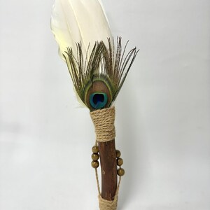 May include: A ceremonial fan featuring a large white feather layered with a peacock eye feather. The handle is a dark brown wooden stick wrapped in tan twine, accented with two strands of small brown wooden beads hanging from the twine wrap.