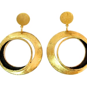 May include: A pair of gold-colored hoop earrings with a circular design. Each earring features a small, round stud at the top and a larger, textured hoop with a black inner section. The earrings are displayed against a white background.