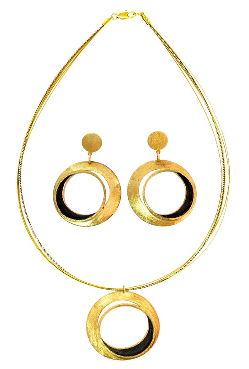 May include: A gold-toned jewelry set featuring a necklace and matching earrings. The necklace has a pendant with a circular design, and the earrings are large hoops. The set has a brushed metal finish with black accents.