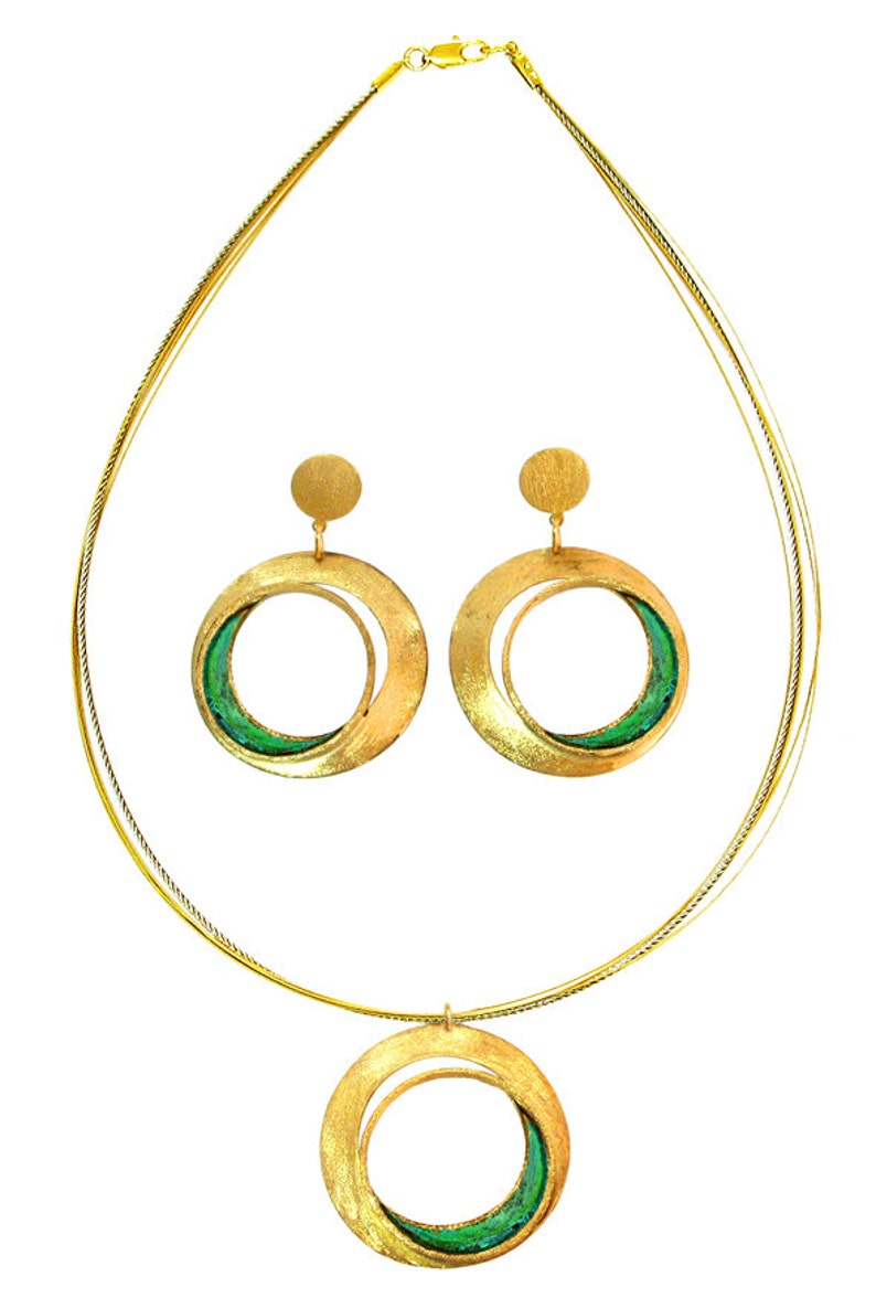 May include: A gold-toned jewelry set featuring a necklace and matching earrings. The necklace has a double-strand chain with a circular pendant. The earrings are also circular. Each piece has a green accent within the circular design.