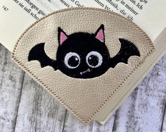May include: A gold leather bookmark with a black embroidered bat with pink ears and white eyes. The bat has a small, white, embroidered smile.