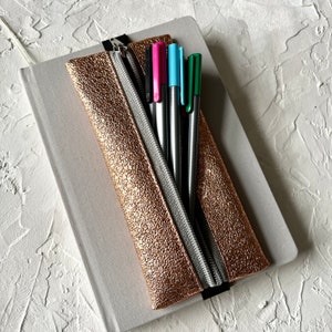 Könnte beinhalten: Eine roségoldene Stiftetui mit Reißverschluss aus Silber und fünf Stiften im Inneren. Die Etui liegt auf einem grauen Notizbuch.