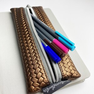 Könnte beinhalten: Eine goldene gewebte Stiftetui mit Reißverschluss. Die Etui ist gefüllt mit bunten Stiften und Markern. Die Etui hat ein schwarzes Etikett mit dem Text "Sweet Surrender".