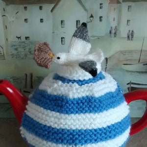 May include: A blue and white striped knitted teapot cosy with a knitted seagull on top. The cosy is made to fit a standard sized teapot.