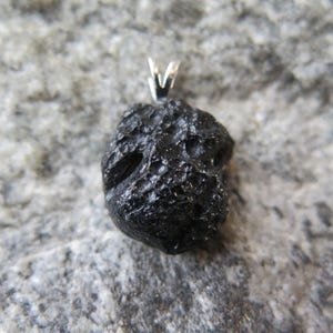 May include: A black pendant with a textured, irregular surface, possibly a meteorite or natural stone. It has a silver-colored bail for a necklace. The pendant is set against a gray, textured background.