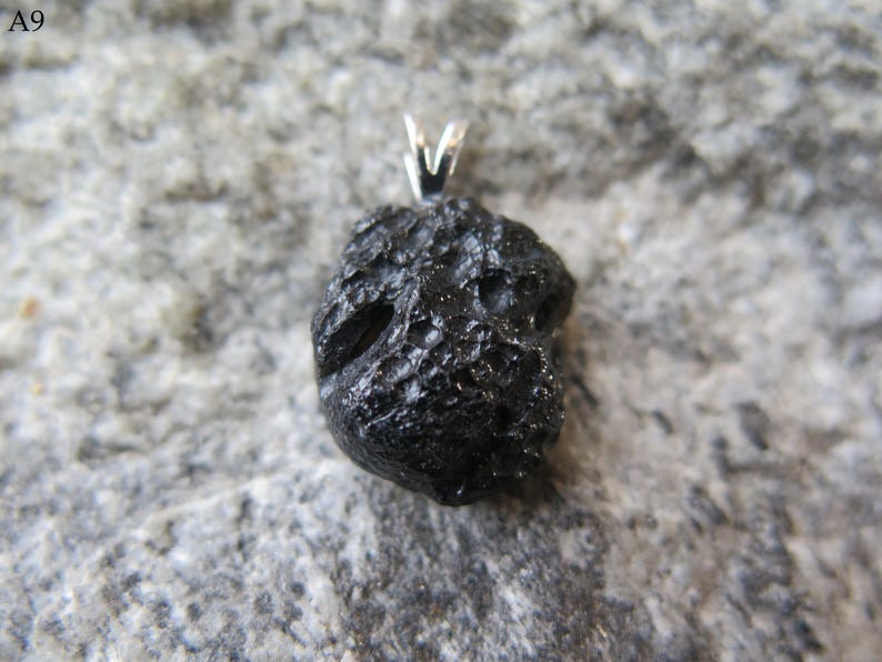 May include: A black pendant with a textured, irregular surface, possibly a meteorite or natural stone. It has a silver-colored bail for a necklace. The pendant is set against a gray, textured background.