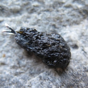 May include: A dark, irregularly shaped pendant with a silver bail. The pendant has a rough, textured surface and is set against a gray, stone-like background. The pendant is a natural stone.