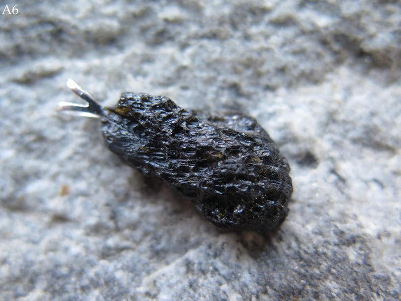 May include: A dark, irregularly shaped pendant with a silver bail. The pendant has a rough, textured surface and is set against a gray, stone-like background. The pendant is a natural stone.