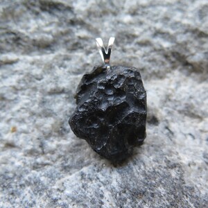 May include: A black, irregularly shaped pendant with a silver bail. The pendant has a rough, textured surface and is set against a gray stone background. The pendant is a unique piece of jewelry.