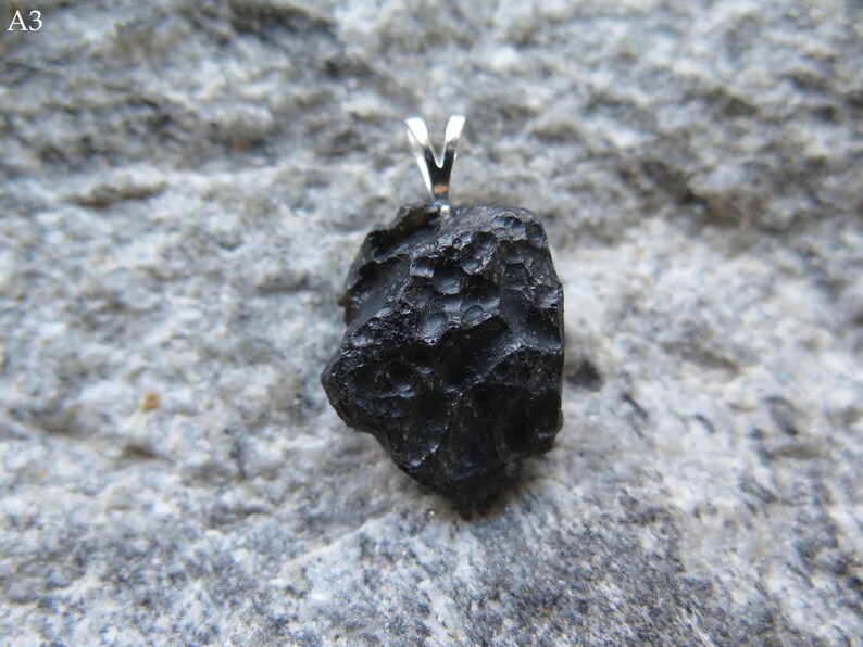 May include: A black, irregularly shaped pendant with a silver bail. The pendant has a rough, textured surface and is set against a gray stone background. The pendant is a unique piece of jewelry.