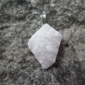 May include: A white, rough-textured, crystal pendant with a silver bail. The pendant is on a gray, textured surface.