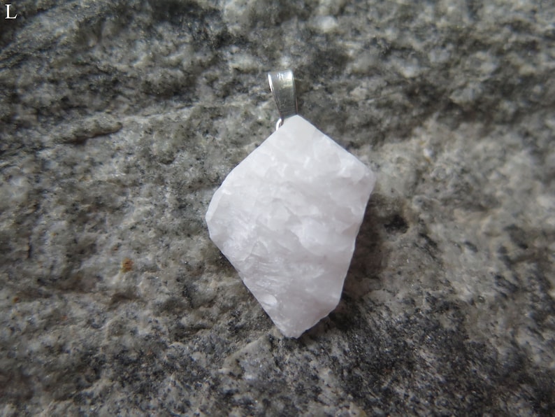 May include: A white, rough-textured, crystal pendant with a silver bail. The pendant is on a gray, textured surface.