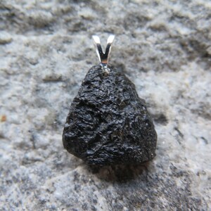 May include: A dark, irregularly shaped pendant with a silver bail. The pendant has a rough, textured surface and is set against a gray, stone-like background. The pendant is a natural stone.