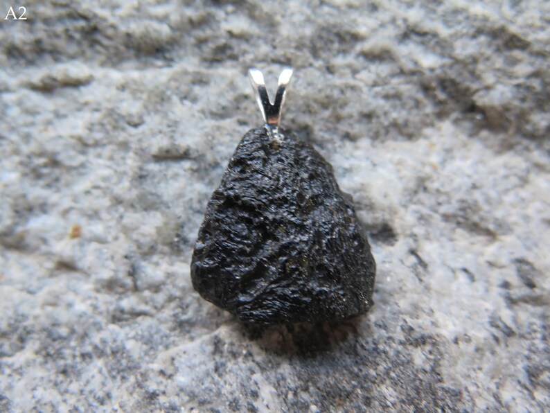 May include: A dark, irregularly shaped pendant with a silver bail. The pendant has a rough, textured surface and is set against a gray, stone-like background. The pendant is a natural stone.