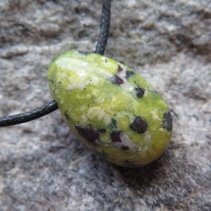 Pode incluir: Um pingente de pedra verde e branca com manchas pretas e castanhas, em um cordão preto. A pedra oval tem um acabamento polido. O fundo é uma superfície cinza texturizada.