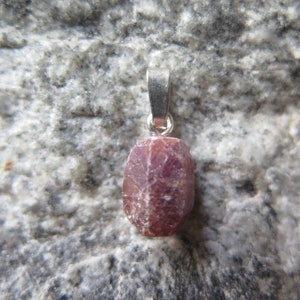Op de afbeelding: Een ruwe robijn edelsteen hanger met een zilveren beugel. De robijn is diep rood van kleur en heeft een ruwe, natuurlijke textuur.