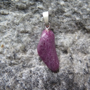 Peut inclure: Un pendentif en pierre de couleur magenta profond, de forme irrégulière et polie, présentant une texture lisse et brillante. La pierre est fixée à une bélière en métal argenté simple sur le dessus, conçue pour être portée en collier.