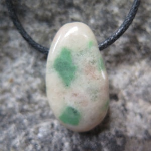 Pode incluir: Um pingente de pedra branca e verde em um cordão preto. A pedra tem formato de lágrima e é salpicada de verde e rosa.