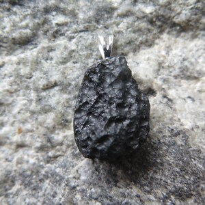 May include: A black pendant with a textured surface and a silver-colored bail. The pendant is a dark, irregular shape, possibly a stone or mineral. The background is a gray, textured surface.