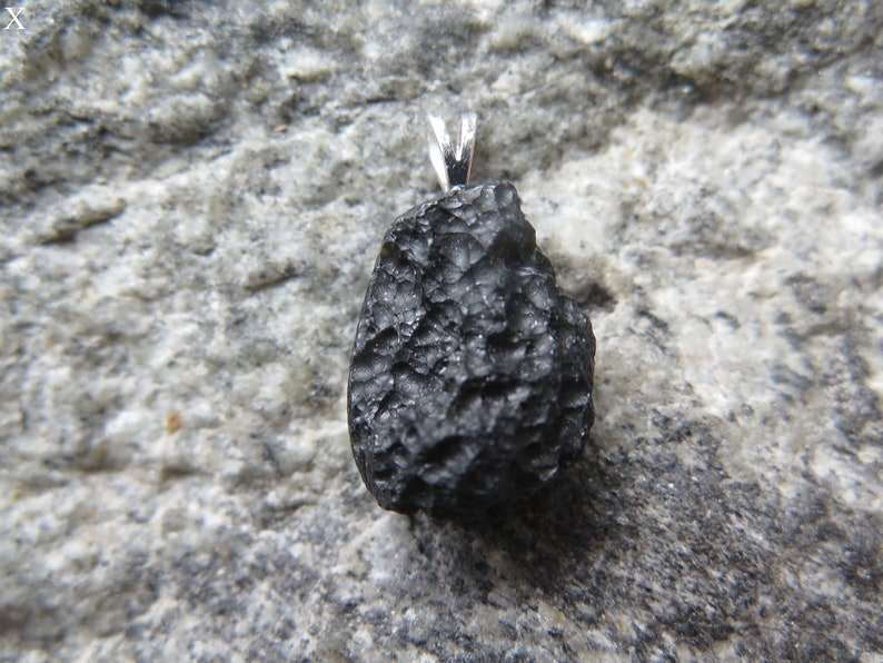 May include: A black pendant with a textured surface and a silver-colored bail. The pendant is a dark, irregular shape, possibly a stone or mineral. The background is a gray, textured surface.