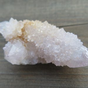 Könnte beinhalten: Ein Cluster aus hellrosa Quarz-Kristallen mit einem blassgelben Farbton an der Spitze. Die Kristalle haben eine raue, strukturierte Oberfläche und sind in einer stacheligen Formation angeordnet. Der Quarz ist vor einem Holzgrund platziert.