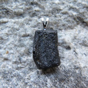 May include: A black, irregularly shaped pendant with a silver bail. The pendant is a rough, natural stone, likely a mineral or crystal. The background is a textured, gray stone surface.