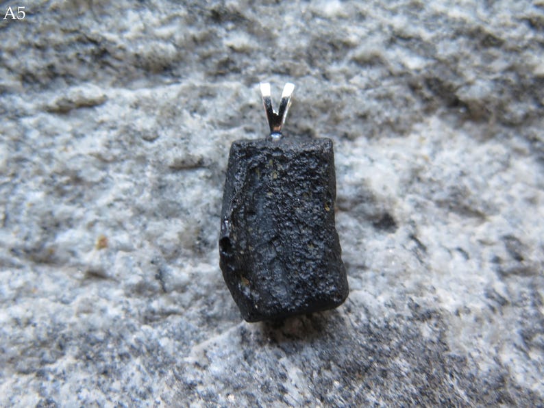 May include: A black, irregularly shaped pendant with a silver bail. The pendant is a rough, natural stone, likely a mineral or crystal. The background is a textured, gray stone surface.