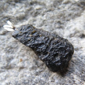 May include: A dark, textured, oblong-shaped stone pendant with a silver-colored bail. The stone is a deep black color and has a rough, uneven surface. The pendant is set against a gray, textured background.