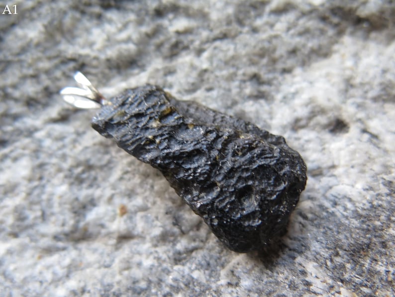 May include: A dark, textured, oblong-shaped stone pendant with a silver-colored bail. The stone is a deep black color and has a rough, uneven surface. The pendant is set against a gray, textured background.
