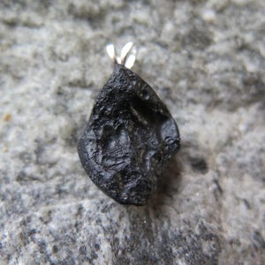May include: A black, irregularly shaped pendant with a silver bail. The pendant has a rough, textured surface and is set against a gray stone background. The text "A7" is in the upper left corner.
