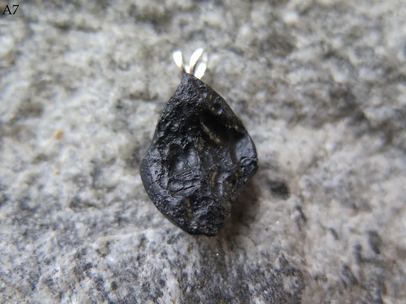 May include: A black, irregularly shaped pendant with a silver bail. The pendant has a rough, textured surface and is set against a gray stone background. The text "A7" is in the upper left corner.