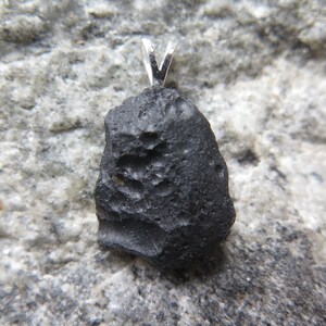May include: A black, irregularly shaped pendant with a silver bail. The pendant has a rough, textured surface and is set against a gray, speckled background. The pendant is likely a natural stone or mineral.
