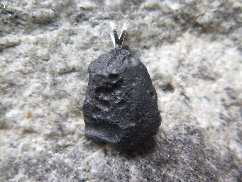 May include: A black, irregularly shaped pendant with a silver bail. The pendant has a rough, textured surface and is set against a gray, speckled background. The pendant is likely a natural stone or mineral.
