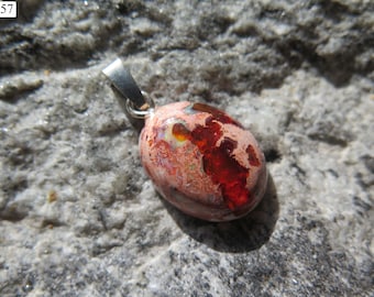 Può includere: Un ciondolo di forma ovale con una pietra di opale lucida e multicolore, caratterizzata da vivaci riflessi rosso intenso, arancioni e iridescenti, montata su una contromaglia in metallo color argento. La pietra presenta una superficie ruvida.