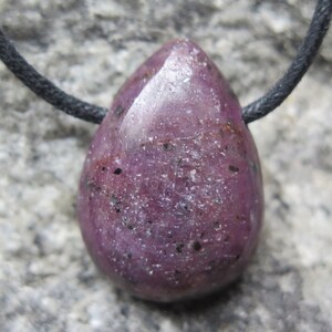Pode incluir: Um pingente de rubi polido em forma de gota em um cordão preto. A pedra preciosa é de um vermelho-púrpura profundo com pequenas inclusões pretas e uma superfície ligeiramente texturizada. O cordão é amarrado no topo.