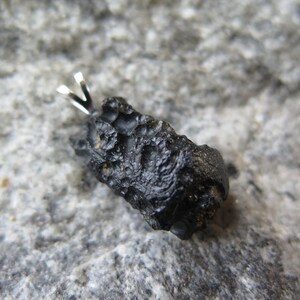 May include: A black, irregularly shaped pendant with a silver bail. The pendant has a rough, textured surface and is set against a gray, speckled background. The text "A10" is in the upper left corner.