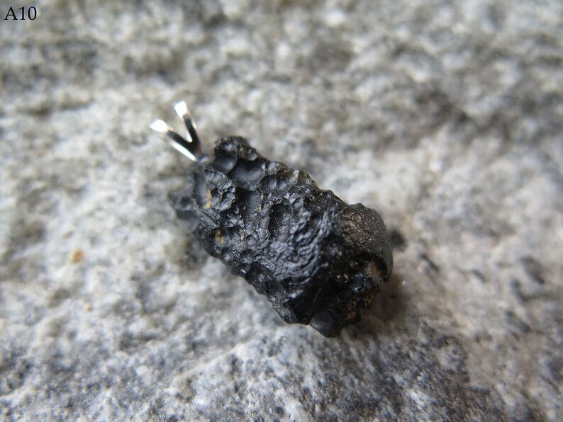 May include: A black, irregularly shaped pendant with a silver bail. The pendant has a rough, textured surface and is set against a gray, speckled background. The text "A10" is in the upper left corner.