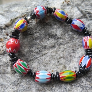 Könnte beinhalten: Ein farbenfrohes Perlenarmband mit ovalen Glasperlen in Rot, Blau, Gelb und Grün mit weißen Akzenten. Die Perlen sind mit schwarzen Abstandshaltern aufgereiht und bilden ein lebendiges und auffälliges Accessoire.