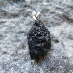 May include: A black pendant with a textured surface and a silver-colored bail. The pendant is irregularly shaped and appears to be a natural stone or mineral. The background is a gray, textured surface.