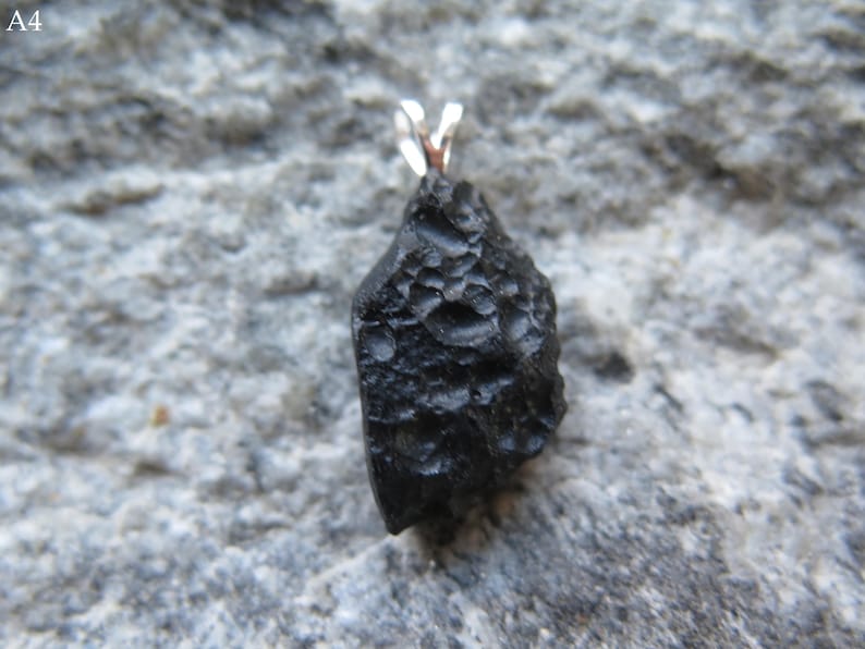 May include: A black pendant with a textured surface and a silver-colored bail. The pendant is irregularly shaped and appears to be a natural stone or mineral. The background is a gray, textured surface.