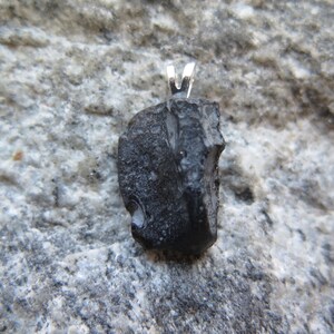 May include: A black pendant with a silver bail. The pendant is a rough, irregularly shaped stone. The stone has a textured surface and appears to be a natural material. The background is a gray stone surface.