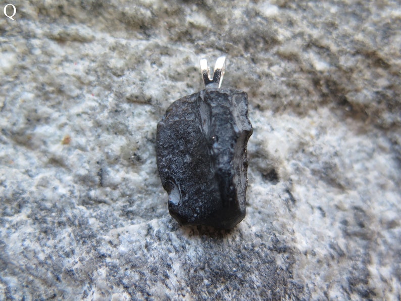May include: A black pendant with a silver bail. The pendant is a rough, irregularly shaped stone. The stone has a textured surface and appears to be a natural material. The background is a gray stone surface.