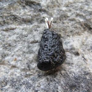 May include: A black, rough-textured pendant with a silver bail. The pendant is irregularly shaped and appears to be a natural stone. The background is a gray, textured rock surface. The pendant is likely a piece of jewelry.