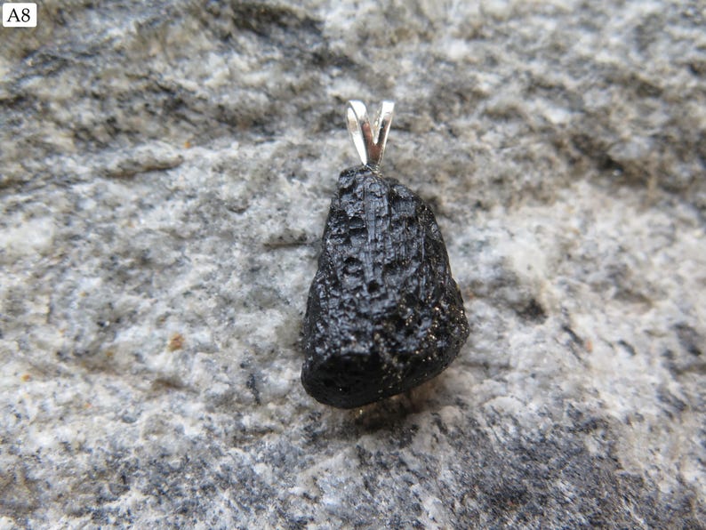 May include: A black, rough-textured pendant with a silver bail. The pendant is irregularly shaped and appears to be a natural stone. The background is a gray, textured rock surface. The pendant is likely a piece of jewelry.
