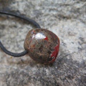 Peut inclure: Une perle en pierre polie et arrondie avec une surface tachetée de brun, de gris et de rouge vif, enfilée sur un cordon noir fin. La perle présente une finition brillante et réfléchissante, avec le texte G10 inscrit dans le coin supérieur gauche.