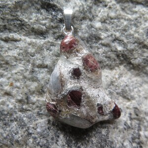 Puede incluir: Un colgante de plata con una piedra gris rugosa con cristales rojos incrustados. La piedra tiene una forma irregular y un aspecto natural y orgánico.