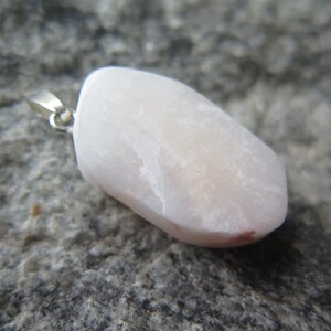 May include: A pale pink, irregularly shaped stone pendant with a silver bail. The stone has a smooth, polished surface and a slightly translucent appearance.