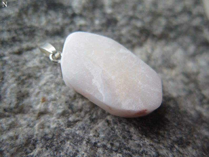 May include: A pale pink, irregularly shaped stone pendant with a silver bail. The stone has a smooth, polished surface and a slightly translucent appearance.